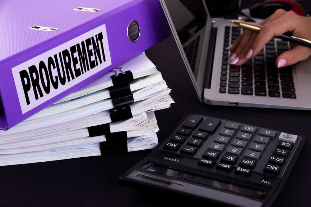 Text, word Procurement is written on a folder lying on documents on an office desk with a laptop and a calculator.